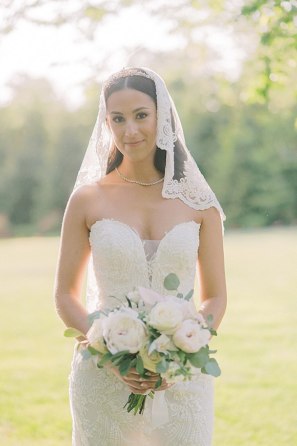 Bridal portrait of a bride holding bouquet with white roses and eucalyptus, wearing a lace veil and beaded headband in a sunlit garden