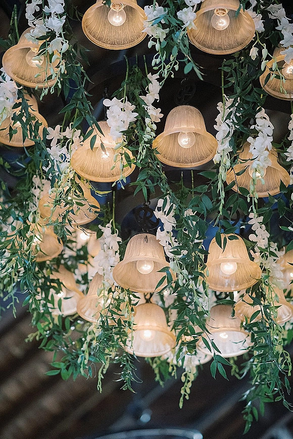 Wedding hanging lights and greenery ceiling installation with exposed bulbs, white flowers, and hanging vines glowing against a dark reception ceiling