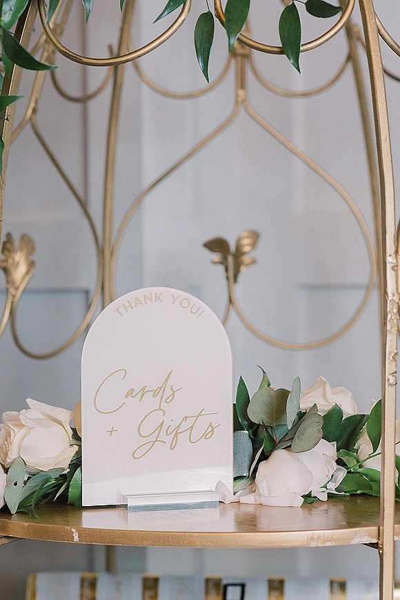 Wedding card table with cards and gifts sign in an acrylic holder, styled with white roses, eucalyptus greenery, and a gold shelf against a light wall