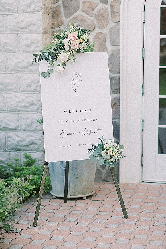 Wedding welcome sign on easel with blush rose and eucalyptus accents beside a stone wall and white door along a paver walkway
