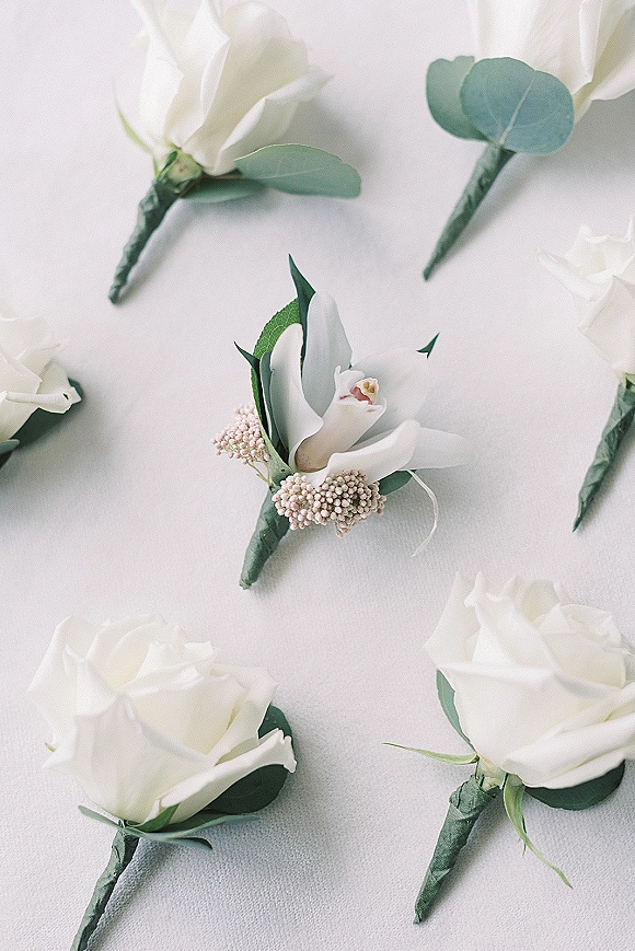 Wedding boutonniere with a white rose, orchid, eucalyptus greenery, and pale pink filler flowers resting on a light fabric surface