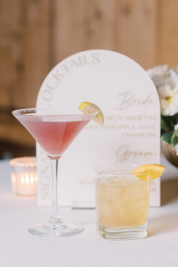 Wedding signature drinks with his and hers cocktails beside an arched menu card, citrus garnishes, and a votive candle on a white tablecloth