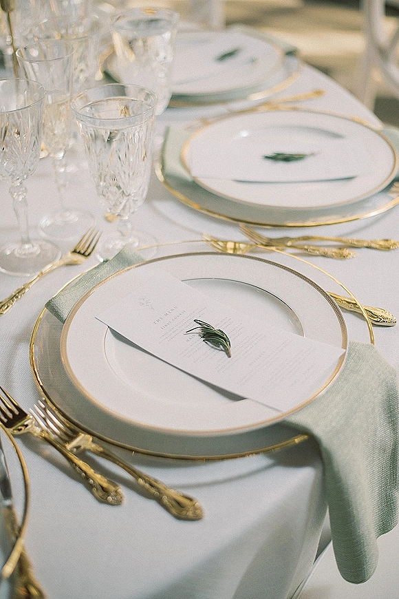 Reception tablescape with wedding place setting featuring gold rim plates, crystal glassware, sage green napkins, and rosemary sprigs in soft indoor light
