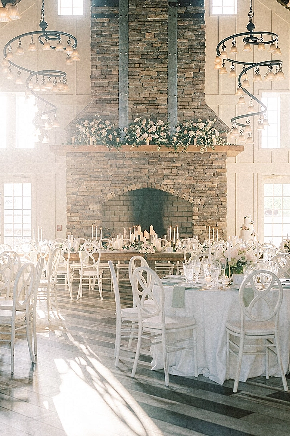 Reception tablescape in a white wedding reception with round tables, floral centerpieces and candles, set by a stone fireplace under chandeliers