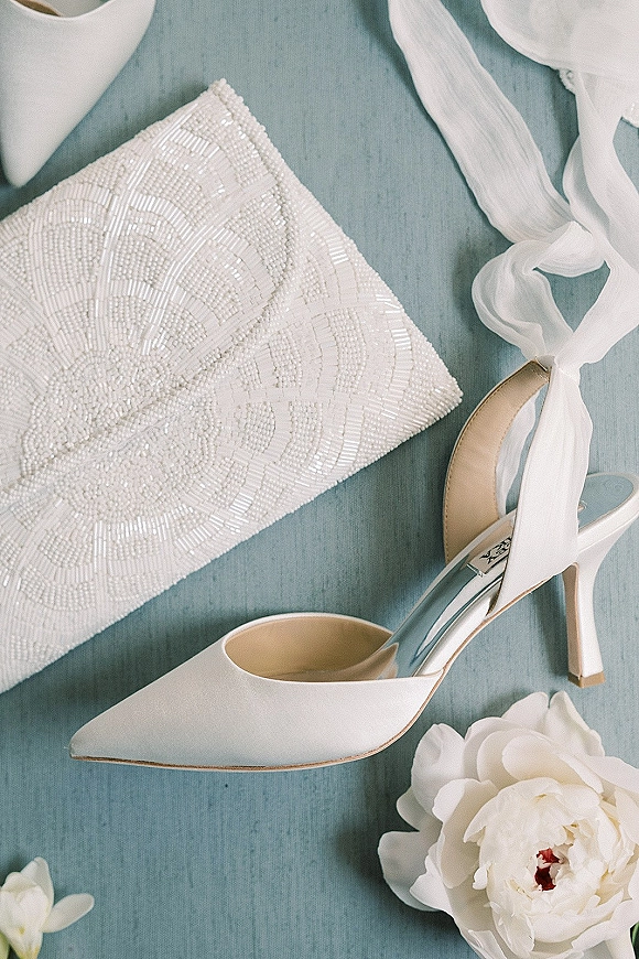 Bridal shoes flatlay with white pointed toe heels and ankle tie ribbon beside a beaded clutch and white peony on blue fabric background