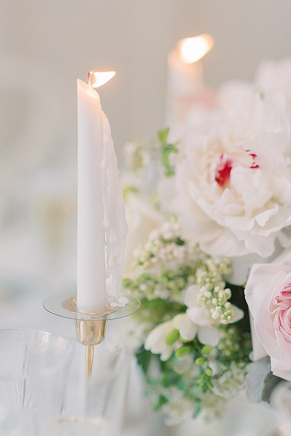 Wedding centerpiece with taper candle centerpiece in a glass holder, dripping wax beside peonies, pink roses, greenery, and a champagne flute on white linen