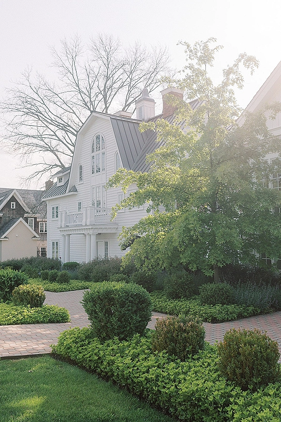 Wedding venue exterior with white clapboard siding, metal roof and balcony, brick walkway through hedges in warm sunlight under trees