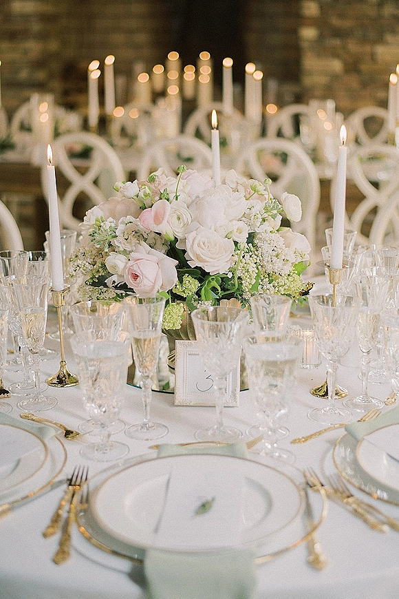 Reception tablescape with wedding table centerpiece of blush roses, white blooms and greenery, taper candles in brass, crystal stemware by brick wall