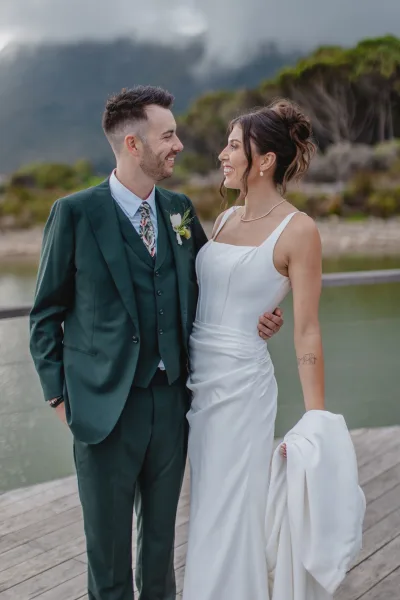Couple portrait of bride in a white dress and groom in a green suit smiling on a wood deck by water with misty mountains behind