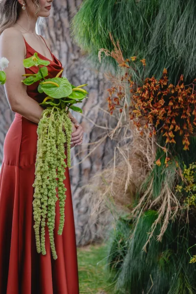Bridesmaid portrait in a rust bridesmaid dress, holding anthurium bouquet with hanging amaranthus, side profile by greenery and tree trunk