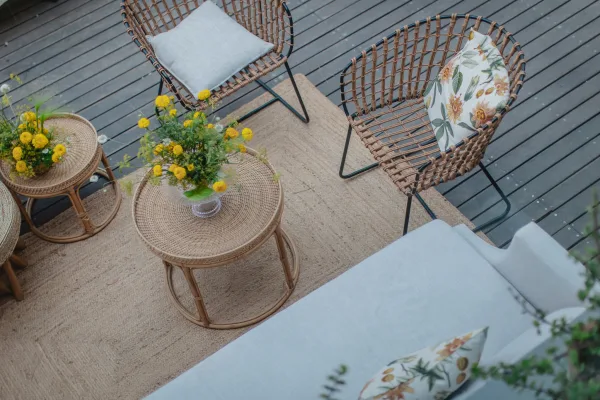 Lounge seating area with woven rattan chairs, jute rug, and sofa with floral pillows around rattan tables and yellow flowers on a deck amid greenery