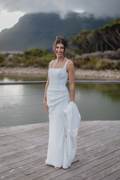 Bridal portrait of a bride in a strapless satin wedding dress holding her train on a wood deck by a lake with mountains under clouds