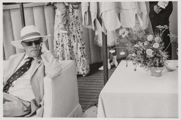 Wedding guest portrait of an older man in a wide brim hat and sunglasses, seated by a table with floral centerpiece on a wood deck