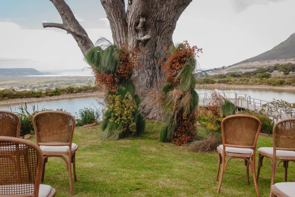 Ceremony altar decor for outdoor ceremony setup with greenery and burnt orange florals on a tree, cane chairs on a lakeside lawn with mountains