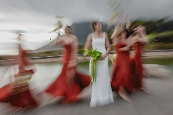 Bride with bridesmaids in red dresses holding bouquets, bride with green bouquet in white gown by a lakeside mountain shoreline under cloudy sky