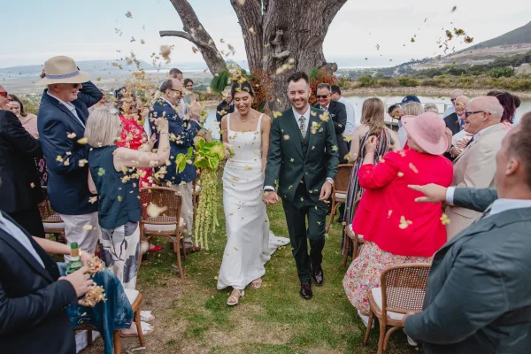 Wedding recessional as newlyweds walk the aisle during their ceremony exit, bride holding bouquet while guests toss confetti by a lakeside lawn