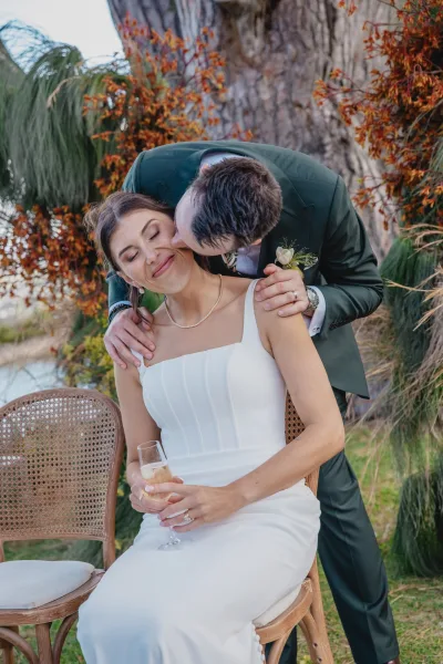 Wedding couple portrait with groom kissing bride’s cheek as she sits holding a champagne flute beside autumn leaves and water backdrop
