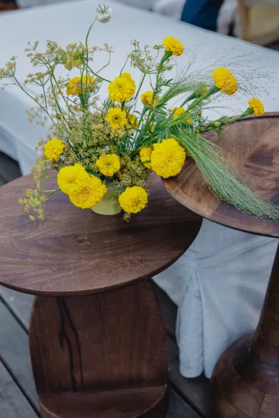 Wedding centerpiece with yellow wedding centerpiece blooms, wildflowers and greenery in a small vase on wooden side tables over a white tablecloth