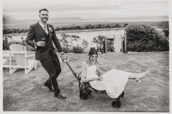 Wedding couple portrait with bride riding in a utility wagon as groom pulls, laughing on a lakefront lawn near dock and Adirondack chairs