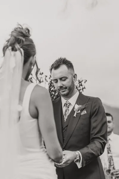 Wedding vows as bride and groom hold hands, groom looking down in an outdoor ceremony with greenery, guests, and veil accent