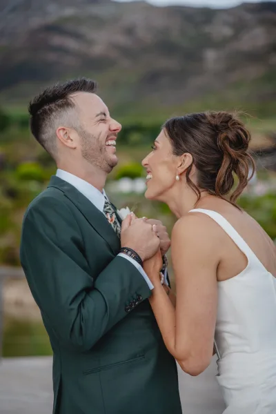 Couple portrait of bride and groom laughing, holding hands with pearl earrings accent, against mountains and stone wall outdoors