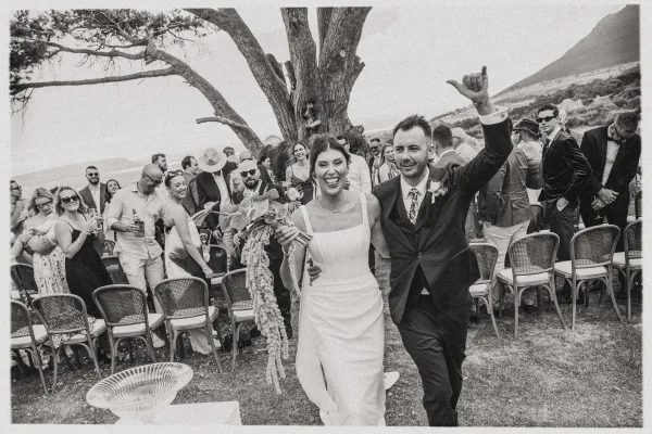 Wedding recessional as newlyweds walking aisle, bride holding bouquet while groom raises a hand past cheering guests under a large tree