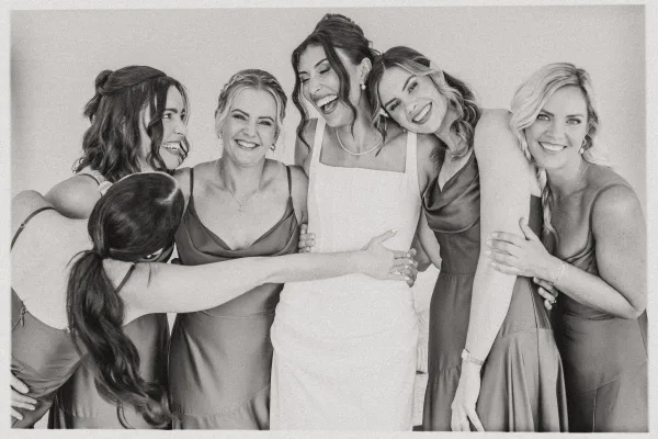 Bridesmaid group photo of bride with bridesmaids hugging her, showing satin dresses and bridal jewelry against a plain wall backdrop