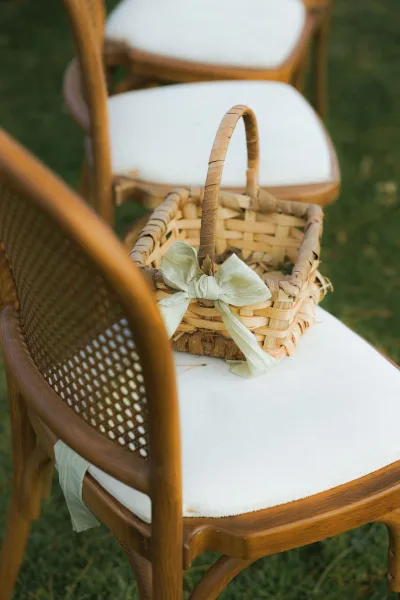 Flower girl basket, woven flower basket with a white bow ribbon on a wooden chair with white cushion, set on a grass lawn