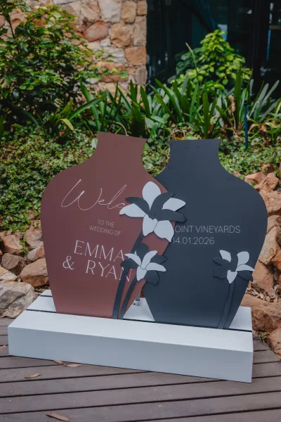 Wedding welcome sign with acrylic wedding welcome sign panels, script lettering, and cutout flower accent, set by garden greenery and stone wall