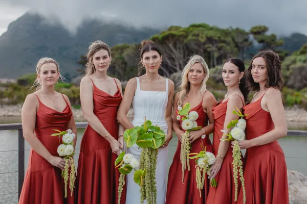Bridal party portrait with bridesmaids with bride in rust dresses, holding white and green bouquets with cascading greenery by a lake and mountains