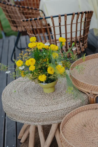 Reception centerpiece with a yellow floral centerpiece in a ceramic vase on a woven side table beside wicker chairs on a wood deck