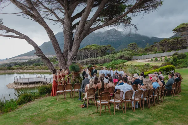 Wedding ceremony outdoors with bride and groom under a large tree by a lake dock, guests in wooden chairs, with bouquet accents