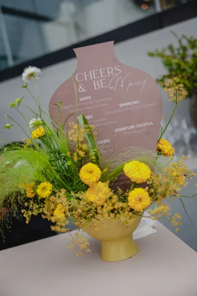 Wedding bar menu and signature cocktail sign on a tabletop with yellow flower bud vase and drinking glasses by modern windowed exterior