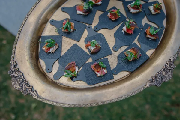Wedding appetizers on black slate serving boards with microgreens garnish and silver tray presentation set over a grass lawn backdrop