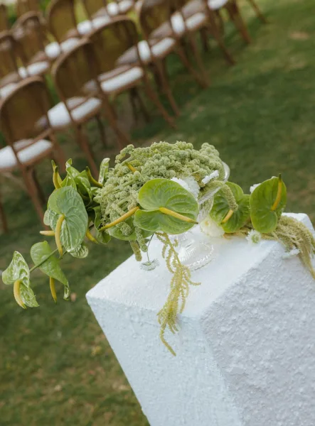 Wedding floral arrangement with green anthurium centerpiece and hanging amaranthus in a glass vase on a textured white pedestal outdoors