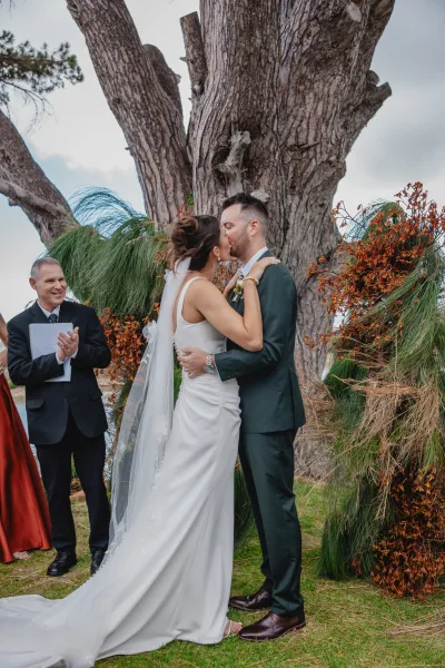 Wedding kiss as bride in white dress and veil kisses groom in green suit beneath greenery arch, officiant holding papers by tree backdrop