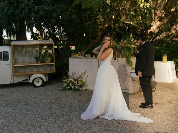 First look moment as bride covers her mouth, reacting to groom in tuxedo by a vintage truck bar with string lights and florals in a garden setting