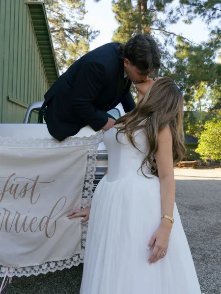 Wedding kiss as groom dips bride in lace-trim dress beside a vintage pickup truck with just married banner, barn and trees behind