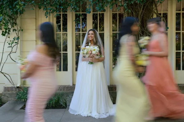 Bridal portrait of a bride holding bouquet in a strapless wedding dress, with blurred bridesmaids passing by near ivy-clad glass doors