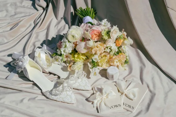 Bridal details flat lay featuring wedding shoes flat lay with lace heels, pearl ankle straps, bouquet, ring box and vow books on satin drape
