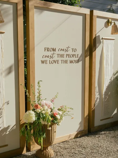 Wedding welcome sign with wood framed wedding sign calligraphy, pastel flowers, and wicker basket against white panels with foliage shadows