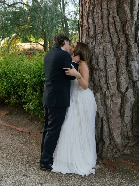 Wedding kiss portrait of bride and groom kissing by a tree, her bracelet visible as they embrace on a gravel garden path with greenery