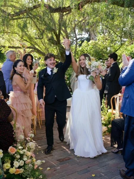 Wedding recessional as newlyweds walk down aisle with raised hands, bride holding bouquet and veil, guests cheering along brick walkway in garden trees