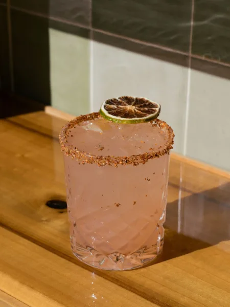 Signature wedding cocktail with ice in a rocks glass, chili salt rim and dried citrus garnish on a wood bar top with tiled wall backdrop