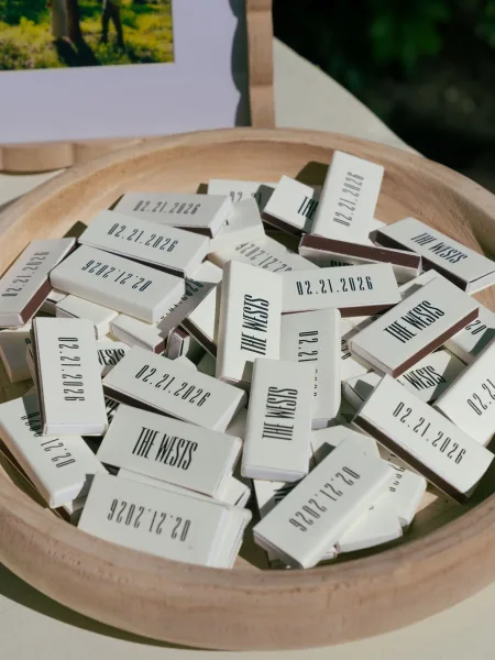 Wedding favors displayed as custom matchboxes wedding in a wooden serving bowl with printed labels on a tabletop with framed photo and greenery