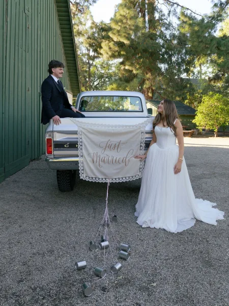 Couple portrait beside a wedding getaway car with a just married sign and tin cans on a vintage pickup by a sunlit barn wall