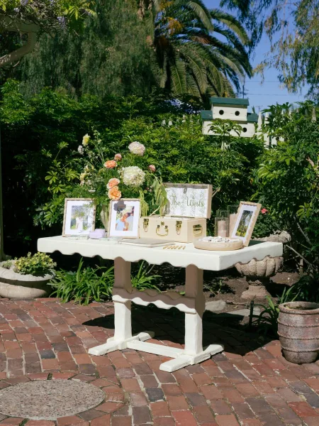 Wedding welcome table with a floral arrangement, framed photos, suitcase card box, guestbook and candles on a brick patio by garden greenery.