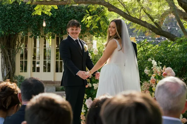 Ceremony moment as bride and groom hold hands under a floral arch, her long veil trailing, with string lights and garden guests behind