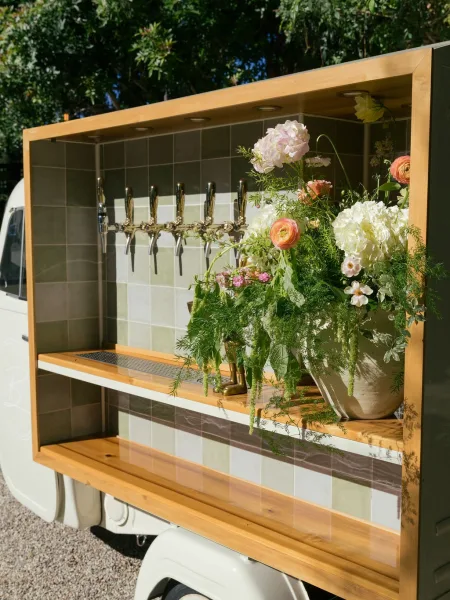 Mobile bar setup with wedding mobile bar beer taps, sage tile backsplash, wood shelves, and floral arrangement on a trailer in trees outdoors