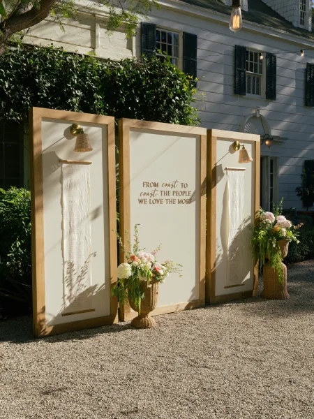Wedding welcome sign with wood framed wedding sign panels and calligraphy, flanked by florals and wicker planters outside a white house with string lights